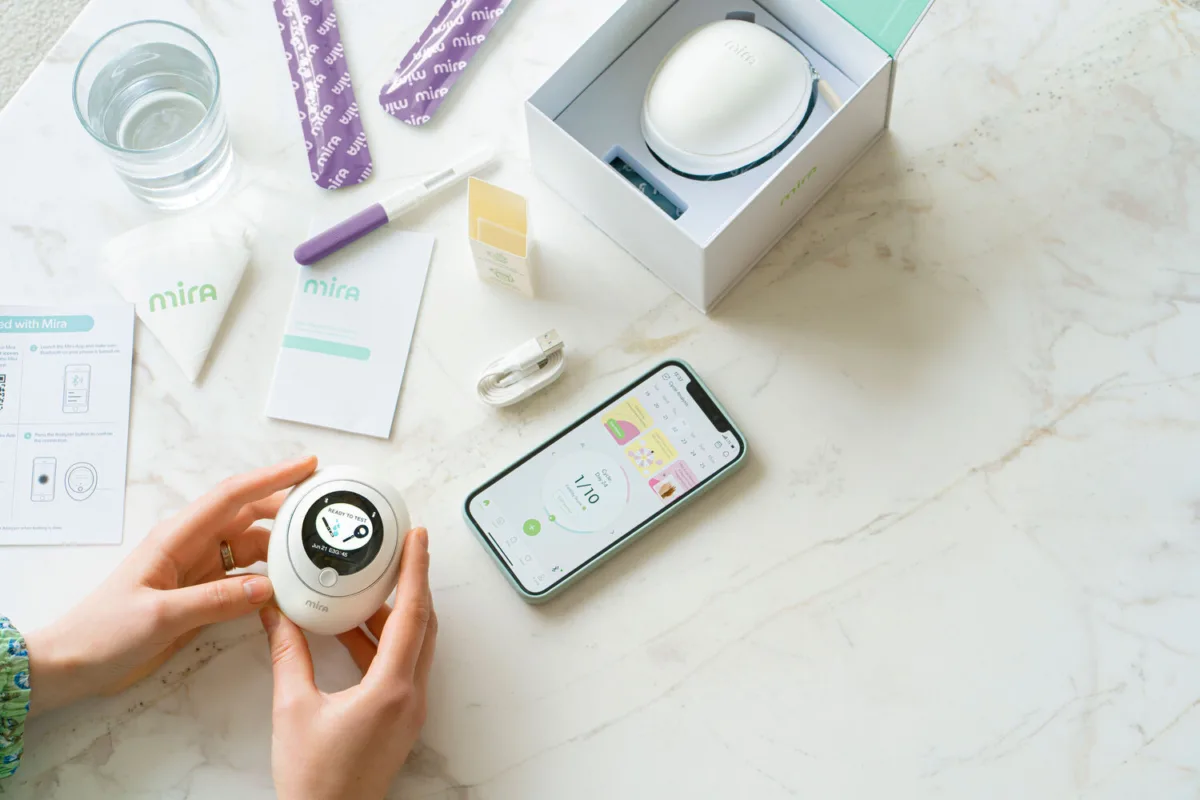 Hands holding the Mira hormone analyzer next to a phone showing the Mira app and test wands